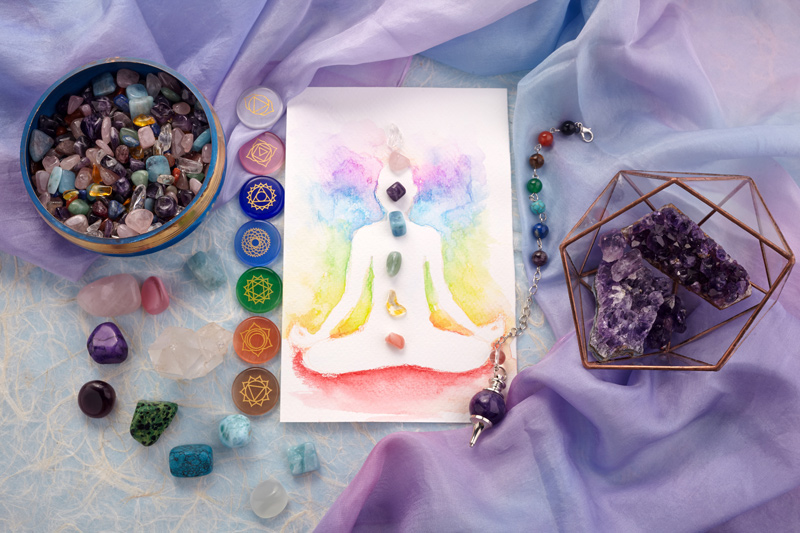 A drawing of a person meditating is covered with gemstones in the 7 chakra colors. Chakra symbols, small gemstones, and an amethyst sit by it on a table.