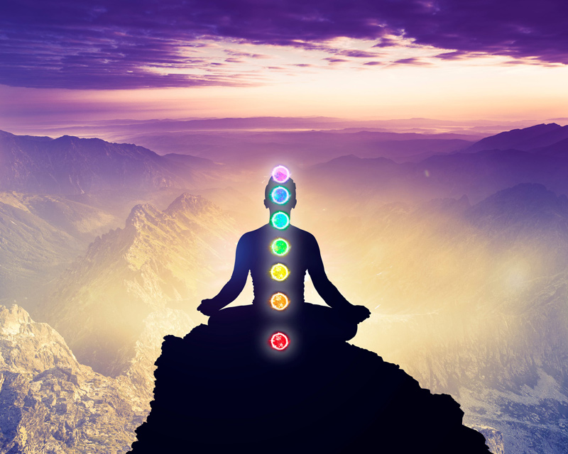 A man is meditating in a yoga pose while sitting on a mountain high in the sky with the 7 colors of the chakras displayed over her body..