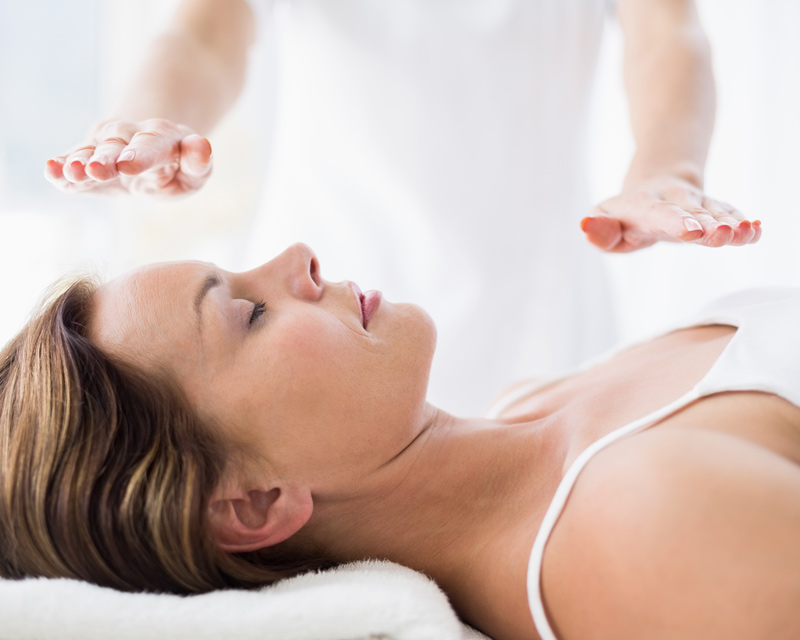A woman lies peacefully on her back while 2 hands are held steadily above her body to perform Reiki Healing. 